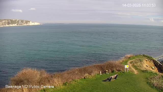 A coastal view shows a calm sea with gentle waves, reflecting a clear sky with some clouds. In the foreground, there is a grassy area with a bench facing the water, surrounded by low bushes. A signpost is visible near the bench, and the cliffs can be seen in the background, leading to a distant shoreline.