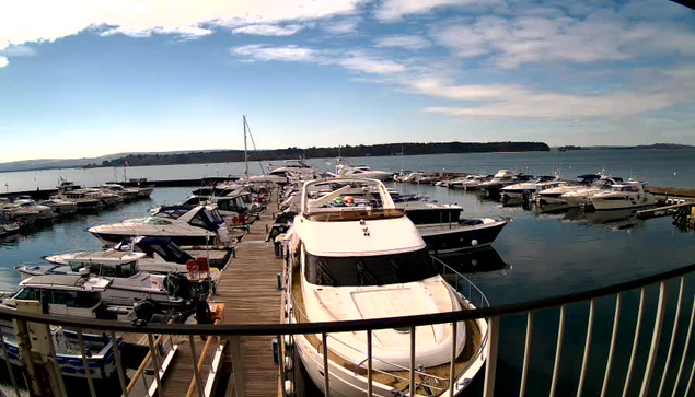 A view of a marina filled with various boats and yachts moored along wooden docks. The water is calm and reflects the sky, which is mostly clear with some clouds. In the distance, a shoreline with trees is visible. The scene captures a serene, sunny day by the water.