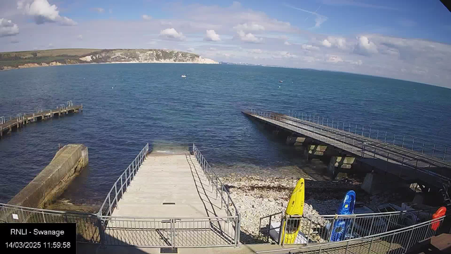 A coastal view with clear blue water and a partially cloudy sky. In the foreground, a concrete ramp leads to the water, bordered by metal railings. To the right, several kayaks in vibrant colors (yellow, blue, and red) are positioned next to the ramp. In the distance, a wooden pier juts out into the water, leading to a curved section. The opposite shore features rolling green hills and chalk cliffs under a bright sky.