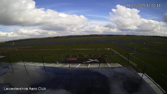 A clear view of an airfield taken from a high vantage point. The foreground shows a flat roof with a railing, and a white 'X' marking on a brown patch. In the distance, there is a grassy airstrip with a few scattered structures and a partially cloudy sky overhead. The scene is well-lit, indicating daytime.