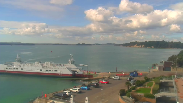 A view of a harbor on a sunny day. A large ferry boat is docked on the left side, with a white and red design. In the foreground, several parked cars are visible, including blue, silver, and red vehicles. To the right, there are small shrubbery and a pathway, along with a few buildings. The water is calm, reflecting the bright blue sky, which features scattered white clouds. Near the horizon, there are small islands.