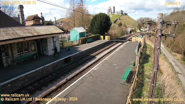 A bright daytime scene of a railway station with stone platform edges. In the foreground, there are several green benches along the platform and a small sign indicating "Way Out." A single person walks towards the station building, which features large windows and a sloping roof. To the left, a blue wooden shed is visible behind a low fence. In the background, a hill rises with the ruins of a castle at its summit, surrounded by trees. The sky is partly cloudy, contributing to a clear and sunny atmosphere. Electric poles line the side of the tracks, which lead off into the distance.