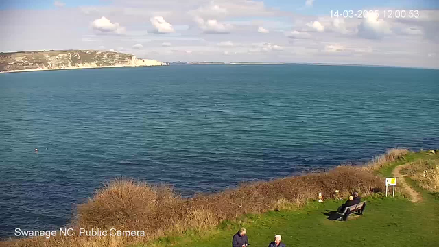 A view of the sea on a sunny day, featuring calm blue waters and a partially cloudy sky. In the foreground, there are two people standing on grass near the edge of the water, while another person is sitting on a bench. In the background, a cliff with greenery is visible along the shoreline. Additional elements include bushes and a signpost near the water's edge.