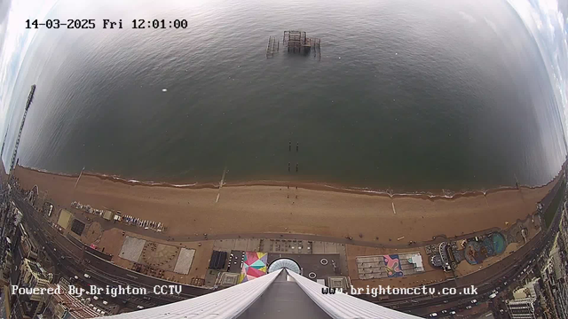 A wide-angle view of a beach scene from a webcam. The image is taken from a height, showing a sandy beach that curves along the shoreline. The ocean appears calm and reflects a cloudy sky. In the middle of the water, there is a partially submerged old pier structure. Along the beach, there are small figures, likely people, walking. On the left side of the image, there are amusement rides and a colorful pattern on the ground near a building. The bottom of the image shows the top of a tower or structure pointing downwards, with a clear view of the beach and the ocean. The date and time are displayed in the top left corner.