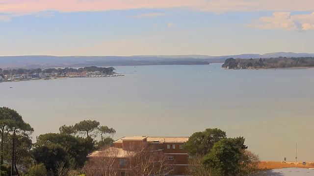 A serene view of a calm body of water under a clear sky. In the background, rolling hills are visible, and a small marina with boats docked is on the left side of the image. The foreground features some trees and buildings, with the water reflecting soft colors from the sky. There are a few boats in the distance, and the scene conveys a peaceful, coastal atmosphere.