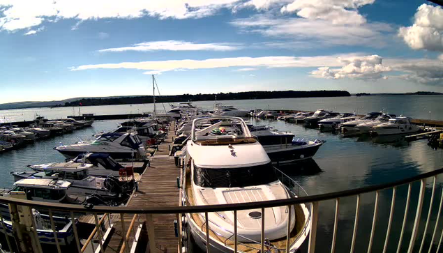 A sunny marina scene with multiple boats docked alongside a wooden pier. The water is calm, reflecting the blue sky and scattered clouds. In the foreground, a large white yacht is moored, while various other boats are visible in the background. The shoreline stretches out in the distance, with green hills and a clear horizon.