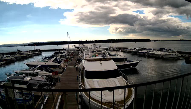 A marina scene featuring numerous boats docked at a wooden pier. Various types of boats, including yachts and smaller vessels, are visible in the water, with some showing colorful covers and equipment. In the background, a cloudy sky transitions to bright areas where sunlight breaks through. The water appears calm with a distant shoreline, partially obscured by trees.