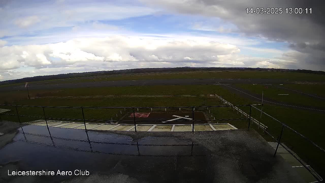 A view from a webcam at Leicestershire Aero Club shows a wide, open landscape under a cloudy sky. In the foreground, there is a railing indicating a balcony or lookout area. Below, a grassy area is visible, with a marked helipad featuring a white "X" design. In the distance, a runway stretches across the scene, bordered by a wooden fence on the left. The ground appears slightly wet, reflecting the surrounding environment. The sky is overcast with varying shades of gray and blue clouds.