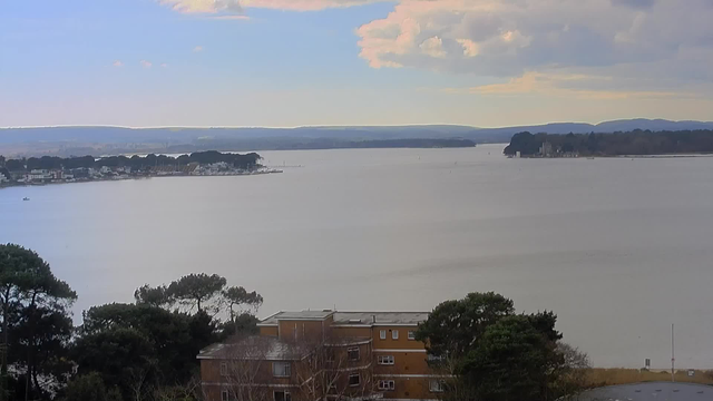 A wide view of a calm lake surrounded by hills and trees. In the foreground, there are a few buildings with a brown exterior and trees. The water reflects the sky, which is partly cloudy with shades of blue and white. In the distance, a shoreline with houses and a marina is visible along the left side of the image. The overall atmosphere is serene and tranquil.