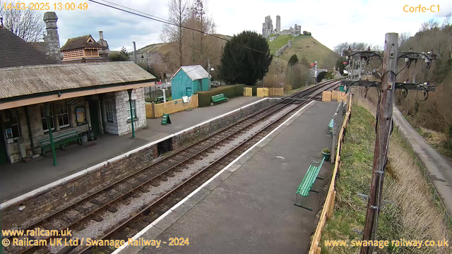 A view of a railway station platform. The station building, made of stone, has a peaked roof and several windows. There are green benches along the platform and a green sign that reads "WAY OUT." Behind the platform, a turquoise shed and a fence made of wooden slats are visible. In the background, there are rolling hills with ruins of a castle on top. The sky is cloudy, and the scene has a tranquil atmosphere with no trains visible on the tracks running beside the platform.