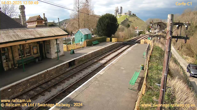 The image shows a railway station with stone and wooden structures. There are two green benches on the platform and a small green shed in the background. The railway tracks run through the center, leading to a hill in the distance upon which a castle ruins are located. The sky is partly cloudy, and there are trees along the sides of the station. A sign indicating "WAY OUT" is visible, and a car is parked along the path beside the station.