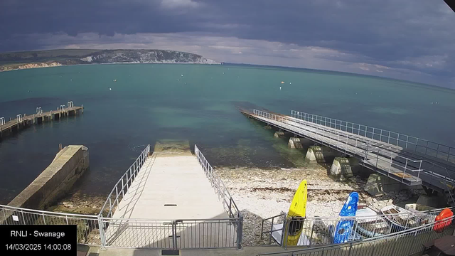 A view of a seaside area with a gray sky and calm turquoise water. In the foreground, there is a concrete ramp leading down to the water, bordered by a metal railing. To the right, two docks extend into the water, one wooden and one metal. Several colorful kayaks, including a yellow and a blue one, are parked on the shore beside the ramp. The background features cliffs and greenery under the overcast sky.