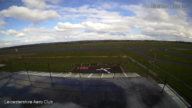 A view from a webcam positioned on a building at the Leicestershire Aero Club. The image captures a wide, open landscape of green grass and asphalt runways under a partly cloudy sky. A large white cross is painted on the runway, positioned centrally in the view. There are several small signs and a fence that runs along the edge of the grassy area. The perspective is from an elevated location, looking out over the aerodrome and the surrounding area.