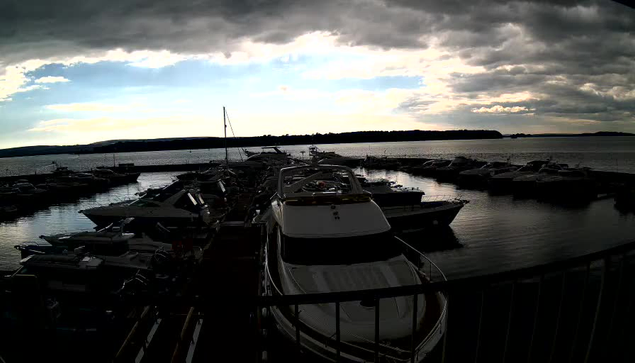 A marina filled with numerous boats docked at the pier. The scene is set under a cloudy sky with a mix of light and shadow cast over the water. In the background, the water reflects the sky, and more boats are visible in the distance. The overall atmosphere appears calm and serene, with hints of blue peeking through the clouds.