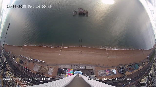 A wide aerial view of a beach and the sea on a cloudy day. The sandy shoreline stretches across the bottom of the image, with gentle waves lapping at it. There is a pier extending into the water on the left side, while an old, partially submerged structure is visible further out in the sea. The beach is lined with a mix of beach activities, possibly including colorful umbrellas and a promenade with buildings. The scene captures a quiet moment at the coastline, with minimal activity on the beach.