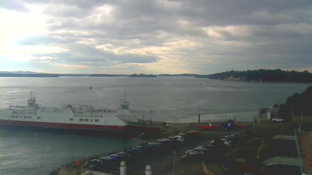 A view of a harbor under cloudy skies. In the foreground, a white and red ferry is docked at the pier, with a sign indicating "Give Way to Chain Ferry" visible on its side. To the right, a parking area filled with cars is visible, and various structures line the waterfront. The water appears calm, reflecting the clouds above, while dark green hills rise in the background.