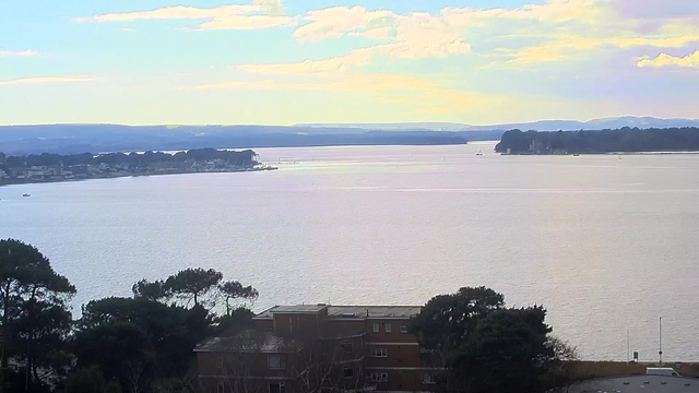 A tranquil view of a wide waterway under a pale sky, with soft clouds scattered overhead. In the foreground, there are patches of greenery from trees along the shore, while a few distant boats are visible on the water. A series of buildings can be seen at the bottom of the image, with a more prominent structure on the left side. The horizon features gentle hills, blending into the sky as it stretches out to the horizon.