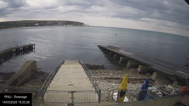 A view of a coastal area with calm water and a cloudy sky. In the foreground, there is a concrete path leading down to the water, flanked by a metal railing. To the right, two boats are visible: one yellow and one blue. At the bottom left, there are pebbles along the shore. In the distance, white cliffs can be seen along the coastline under a grey sky. The scene conveys a tranquil yet overcast atmosphere.