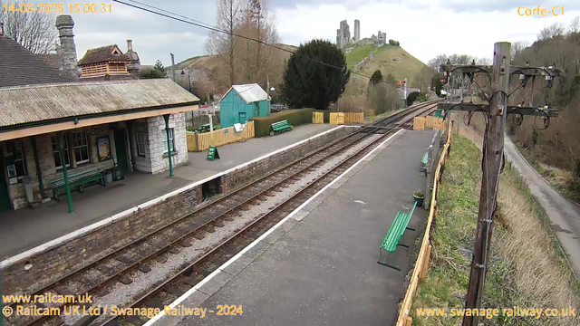 The image shows a country train station with stone and wooden structures. There is a platform with several green benches for seating. To the right, railway tracks stretch into the distance. A wooden utility pole stands on the right side of the image, with electrical wires attached. In the background, a grassy hill is visible, featuring ruins of a castle. There are trees and a small shed near the platform. The sky is overcast with occasional clouds. A sign on the ground reads "WAY OUT."