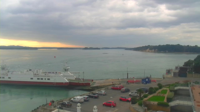 A tranquil waterfront scene showing a bay under a cloudy sky. In the foreground, several parked cars are visible in a lot next to a building with white railings. To the left, a ferry boat is docked at a pier, with its white hull and red lower section standing out against the calm water. There are gentle waves on the surface of the water, reflecting muted colors from the overcast sky. In the background, a mix of green trees and sandy cliffs frame the bay, while a faint outline of hills is seen on the horizon.