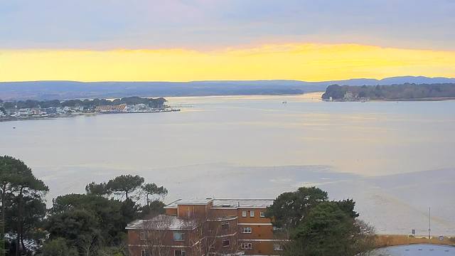 A panoramic view of a calm waterway surrounded by low hills, with a soft gradient of colors in the sky transitioning from pale yellow to light blue. In the foreground, there are several buildings with red-brown facades and greenery, while small boats can be seen on the water in the distance. The landscape is tranquil, conveying a sense of serenity.