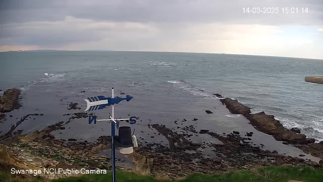 A coastal view showing the sea with waves gently rolling in, against a backdrop of a cloudy sky. In the foreground, there is a weather vane with a blue arrow pointing north and a circular sensor below it. Rocky outcrops are visible in the water, and there is green grass along the edge of the cliff where the camera is positioned. The scene appears calm with no boats or people present.