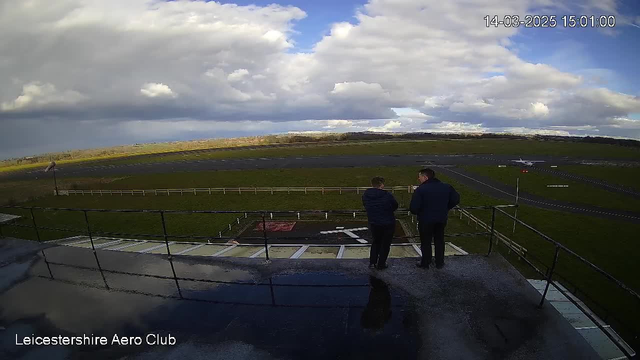 Two men stand on a balcony overlooking a grassy airfield. They face towards the runway, which is marked with painted lines. In the background, a light aircraft is positioned on the runway, with a cloudy sky above. The landscape features a mix of grass and bare earth, and there are scattered clouds. The scene is bright, with some sunlight illuminating the area, and water reflects light on the balcony surface.