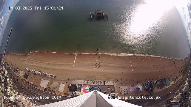 A high-angle view of a beach with light brown sand and a blue ocean, taken from a tall structure. In the distance, a ruined pier is visible jutting out into the water. A few people can be seen walking along the beach. The shoreline is lined with various colorful structures and rides, including a circular blue pool. The sky is clear with a few clouds and sunlight reflecting on the ocean surface. The image includes a timestamp indicating the date and time of the capture.