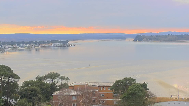 A tranquil seascape at sunset, featuring calm waters reflecting shades of orange and purple in the sky. In the foreground, there are low buildings with vegetation surrounding them. To the left, a waterfront area with boats and piers is visible, while the horizon showcases distant landmasses and trees, creating a serene atmosphere.