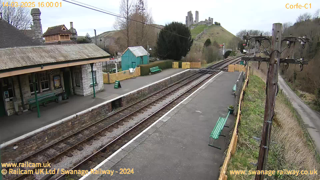 A view of Corfe railway station featuring a stone building with a sloped roof, several green benches along the platform, and a blue shed nearby. The railway tracks run alongside the platform, leading towards hills in the background topped with a castle ruin. Surrounding the station are trees, grass, and a fenced area. In the foreground, a wooden utility pole holds electrical wires. The sky is overcast, giving a grayish appearance to the scene.