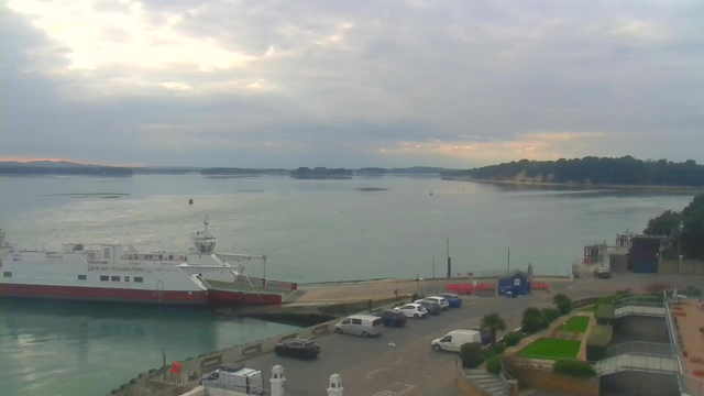 A panoramic view of a harbor under a cloudy sky. In the foreground, a white and red ferry boat is docked at a pier. Nearby, several parked vehicles are visible on a paved area. In the background, the calm water reflects the overcast sky, with gentle ripples and a few distant islands. Several boats are seen in the water, and a small section of greenery and landscaping is visible along the harbor.