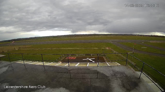 A cloudy sky dominates the image, with patches of light breaking through. Below, an airstrip is visible, bordered by green grass and a wooden fence. In the foreground, there is a helipad marked with a white cross and a red square. The Loughborough Ayr Club's building is in the lower left corner, where the surface appears wet. Several markers and lines are present on the runway, indicating landing paths.