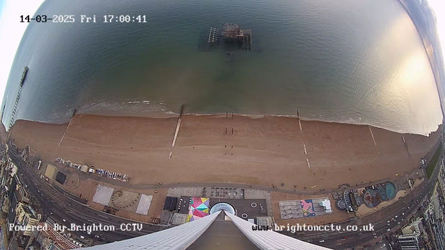 An aerial view of a beach with golden sand and waves gently lapping at the shore. In the distance, an old wooden pier extends into the water, partially submerged. Several individuals can be seen walking along the beach, and colorful structures, including a circular pattern, are positioned near the shoreline. The scene is set against a serene water backdrop with a calm surface reflecting soft light from above. The image is timestamped March 14, 2025, at 5:00 PM.