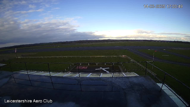 A view from a webcam at Leicestershire Aero Club, capturing a large open field with a runway extending into the distance. In the foreground, there is a flat rooftop area with a railing. A white cross-shaped landing area is visible, surrounded by green grass and a fenced perimeter. The sky is a mix of blue and gray, with scattered clouds.