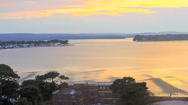 The image shows a scenic view of a calm body of water at sunset, reflecting shades of orange, yellow, and soft blue in the sky. In the foreground, there are trees and a building with a flat roof. In the middle distance, a small marina is visible with boats lined up. The horizon features low hills that fade into the distance, providing a tranquil and picturesque landscape.