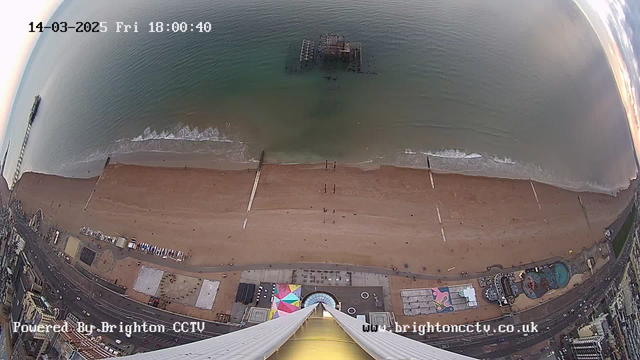 Aerial view of a beach with a sandy shoreline and the ocean. Distinct sand patterns can be seen on the beach, along with groups of people walking. In the distance, an abandoned pier extends into the water. The water is calm with gentle waves, reflecting soft light. Colorful structures and a boardwalk are visible along the beach, while some amusement attractions are seen near the shoreline. The image is timestamped to show the date and time.