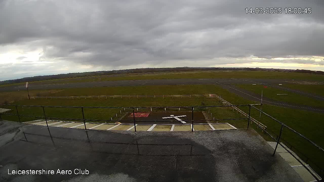 A cloudy sky is visible above an open field with a runway at the Leicestershire Aero Club. The ground is mostly green, with some dark gravel markings. A white airplane symbol is painted on the ground, near a red square area. A wooden fence borders the runway, with a small flag visible in the distance. The scene is captured from a high vantage point, showing the expansive outdoor space and the overall cloudy atmosphere.