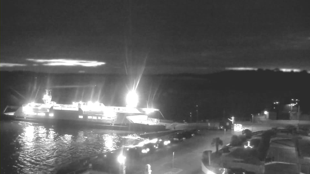 A dark scene shows a ferry docked at a harbor, illuminated by bright lights on its upper deck. The water reflects the lights, creating a shimmering effect. In the background, the sky is dark with hints of clouds and silhouettes of distant hills against the fading light. Vehicles are parked along the waterfront, and additional faint lights are visible in the background. The overall atmosphere conveys a quiet, nighttime setting.