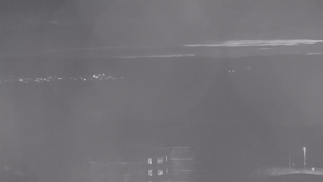 A nighttime view featuring a faint skyline with scattered lights from buildings in the distance, set against dark clouds and an overcast sky. The image has a monochromatic appearance with varying shades of gray, creating a soft, blurred effect. In the foreground, the outlines of a building and streetlight are visible, but details are minimal due to low lighting.