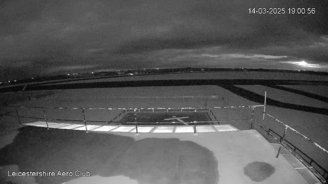 A dark, cloudy sky is visible over a landscape with a runway and a helipad in the foreground. The scene is illuminated by dim lights along the horizon, indicating distant structures. A railing borders the helipad, and the overall image has a stark, monochromatic appearance due to low lighting. The timestamp in the corner indicates the date and time.