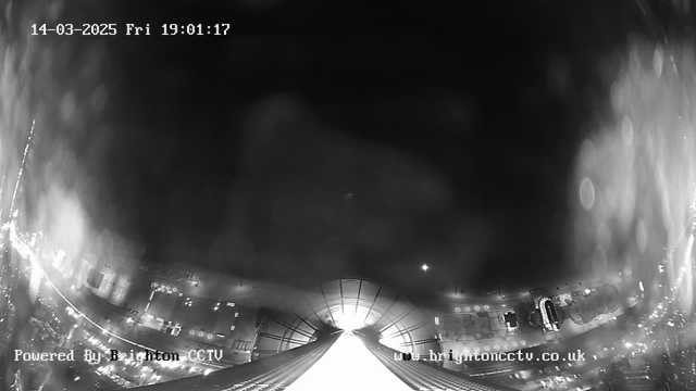 A black and white webcam image showing a wide view of a city street at night, with lights visible that suggest movement. The foreground features a downward slope, possibly a roof or structure leading toward the bottom of the image. The upper part has a curved ceiling with light coming from the center. The time and date are displayed in the top left corner as "14-03-2025 Fri 19:01:17" and the text "Powered By Brighton CCTV" is at the bottom.
