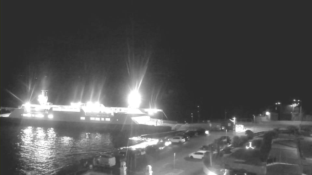 A ferry is docked at a pier, illuminated by bright lights, reflecting on the water’s surface. In the foreground, several parked cars line the waterfront, while the area is surrounded by dark silhouettes of buildings and trees against a night sky.