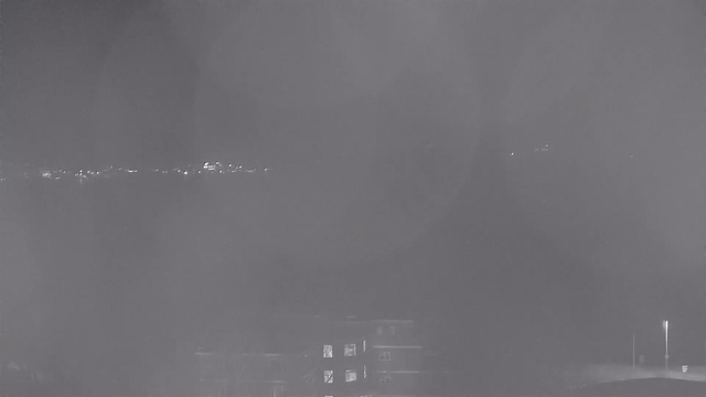 A dimly lit scene captured from a webcam, showing a landscape with a few faint lights in the distance against an overcast sky. In the foreground, there are blurred outlines of buildings, with only a few illuminated windows visible. The overall image appears hazy and lacks detail, suggesting nighttime or low visibility conditions.