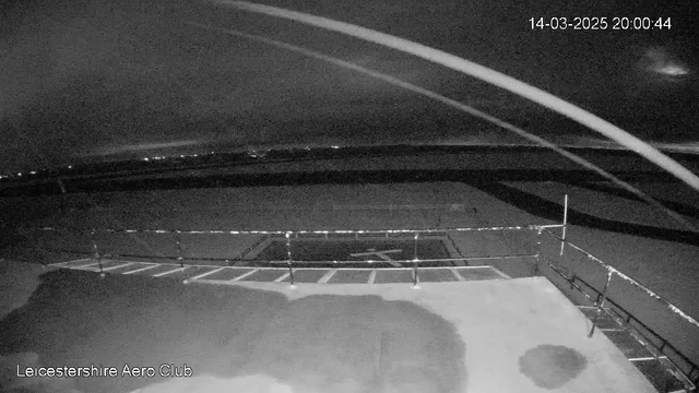 A dimly lit view from a webcam showing an airfield at night. In the foreground, there is a flat surface with a faint outline of a helicopter landing pad marked with a cross. A low barrier fence runs along the edge. The background features a dark sky with some lighter patches and distant lights suggesting a city or town in the far distance. The overall scene is predominantly dark, with only minimal illumination. A date and time stamp is visible in the upper right corner.