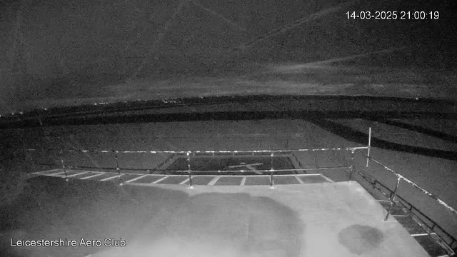 A dimly lit, nighttime image showing an empty tarmac area with a boundary railing on the right side. The background features a dark sky with faint clouds, and a distant glow of lights from the horizon. The tarmac has a marked area for landing, but no aircraft is present. The timestamp at the top indicates the date and time.