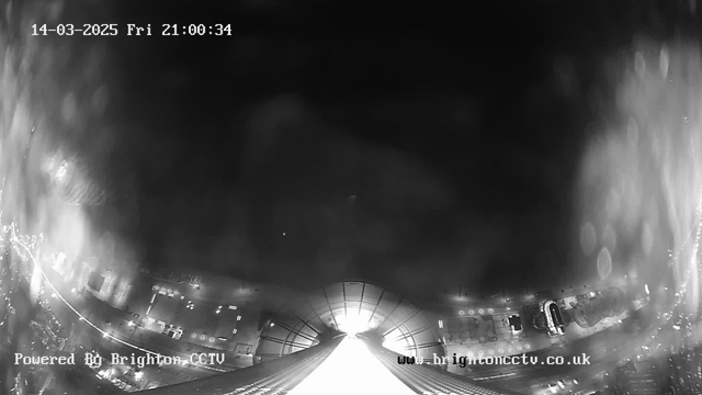 A black and white webcam image showing a view of a wide street at night. The scene is illuminated by bright lights and reflections, with a smooth, illuminated pathway leading toward a distant structure. The top of the image displays the date and time: March 14, 2025, at 21:00:34. In the bottom left corner, there's a watermark indicating it is powered by Brighton CCTV. The overall atmosphere is moody and slightly blurry, suggesting low visibility conditions.