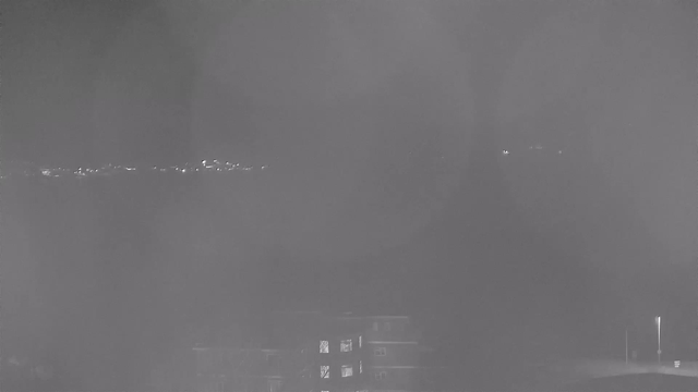 A nighttime webcam image showing a blurred landscape. In the background, faint lights along the horizon indicate a distant city. The foreground features an indistinct building with lit windows. The overall scene is hazy, with a dark ambiance and minimal details visible.