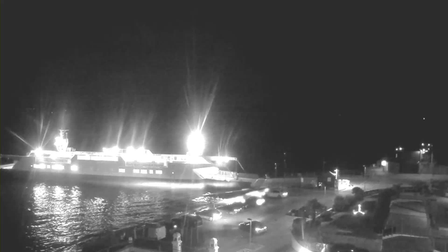 A nighttime scene featuring a brightly lit ferry docked near the shore. The harbor is visible with dark water reflecting the lights. Several cars are moving along the road adjacent to the dock, while light sources illuminate parts of the area. The background is mostly dark with some faint lighting from distant structures.