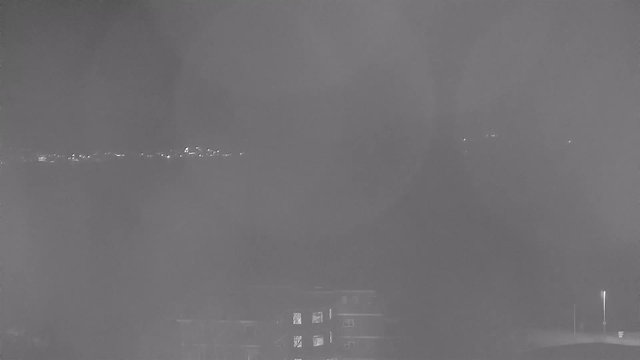 A dimly lit view from a webcam, showing the silhouette of a building in the foreground. In the background, faint lights can be seen along a horizon, resembling distant streetlights or reflections on water. The overall appearance is hazy and monochromatic, with a grayish tint dominating the scene.