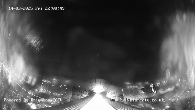 A black and white image from a webcam, showing a low-angle view of a bright light in the distance resembling an entrance or an exit, with blurred lines and abstract shapes surrounding it. The foreground includes the outlines of buildings or structures. The upper part of the image is darker, while the lower area features brighter points of light, suggesting it is possibly a nighttime scene in an urban setting. The date and time in the top left corner indicate it is March 14, 2025, at 22:00:49. The bottom left corner has a text label indicating "Powered By Brighton CCTV."